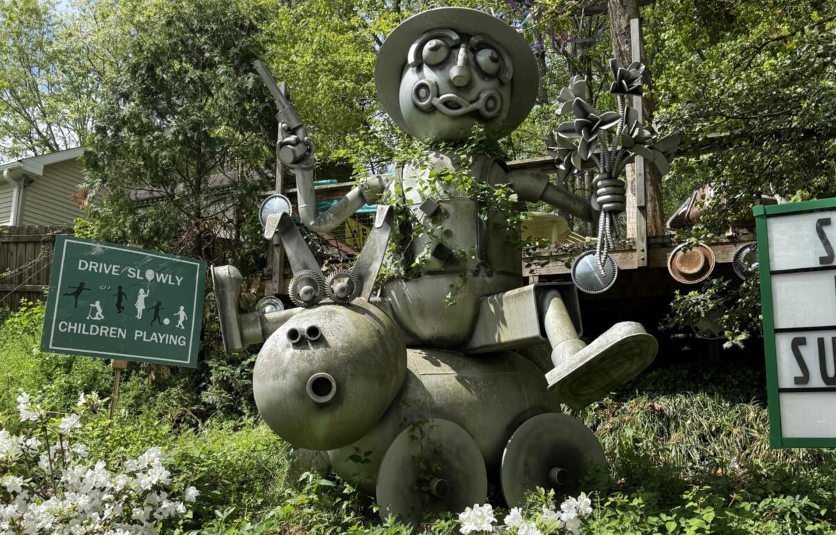 A large metal sculpture depicting a man riding a wheeled pig or dog while carrying a six-shooter and a bouquet of flowers stands at the entrance to the Burton Street community's Peace Gardens & Market.