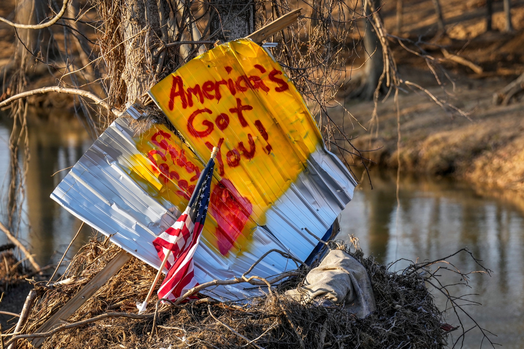 An American flag stands next to a sign written on housing debris along the Swannanoa River, Wednesday, Feb. 5, 2025, in Swannanoa, N.C. Swannanoa was one of several communities in western North Carolina that was severely affected by Hurricane Helene. (AP Photo/Kathy Kmonicek)