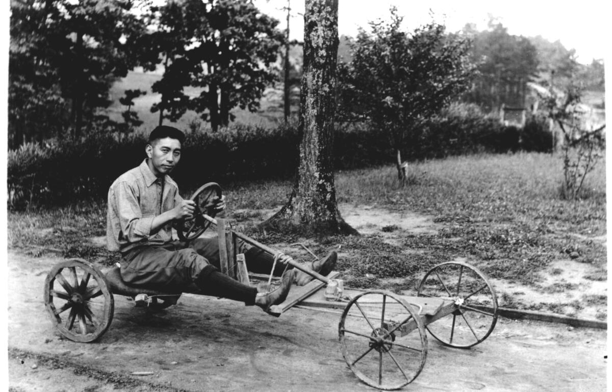Black-and-white photo of a man sitting on a rustic, four-wheeled wooden cart with a steering wheel in a park with trees.