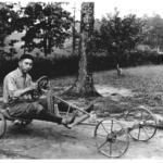 Black-and-white photo of a man sitting on a rustic, four-wheeled wooden cart with a steering wheel in a park with trees.