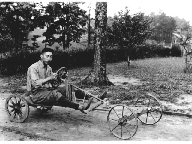 Black-and-white photo of a man sitting on a rustic, four-wheeled wooden cart with a steering wheel in a park with trees.