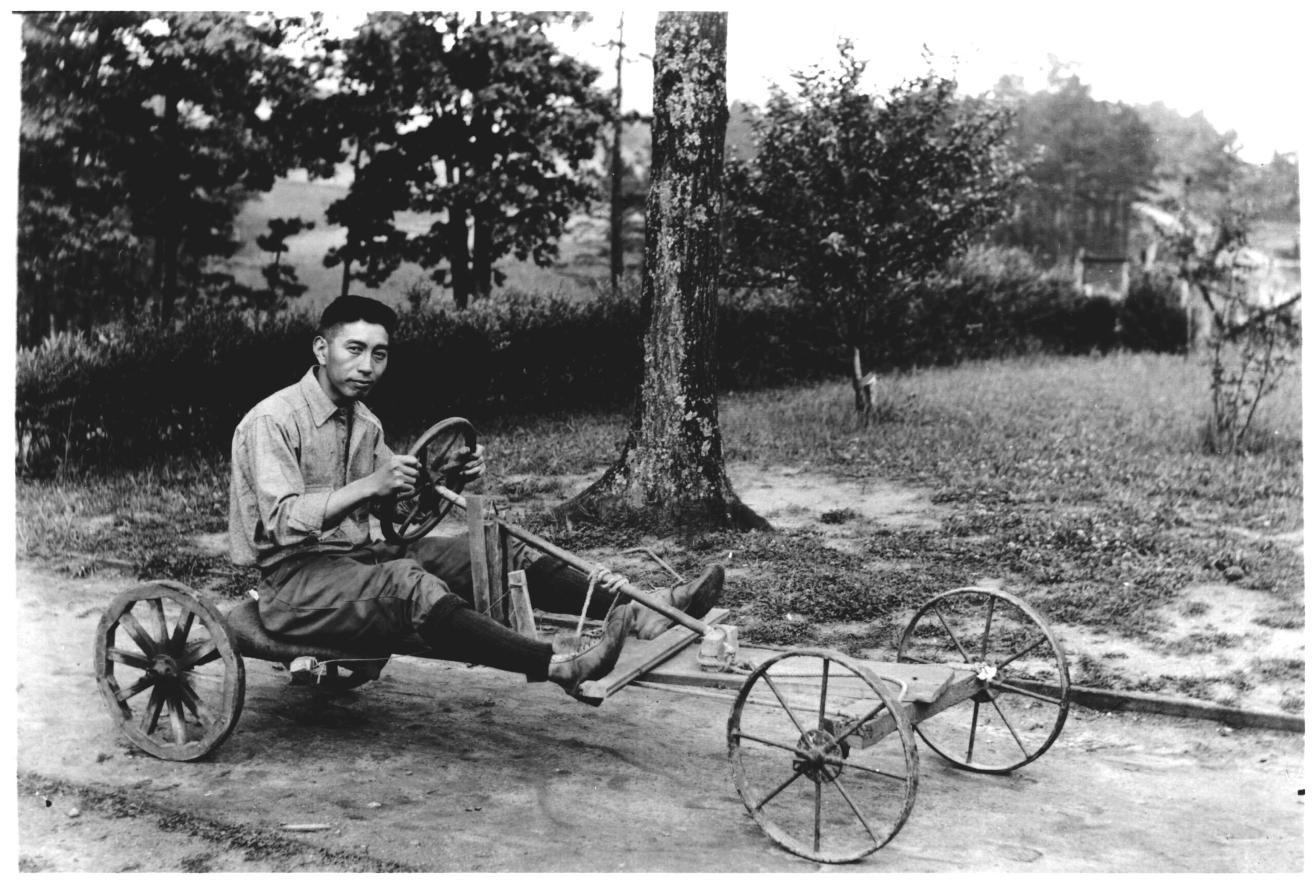 Black-and-white photo of a man sitting on a rustic, four-wheeled wooden cart with a steering wheel in a park with trees.