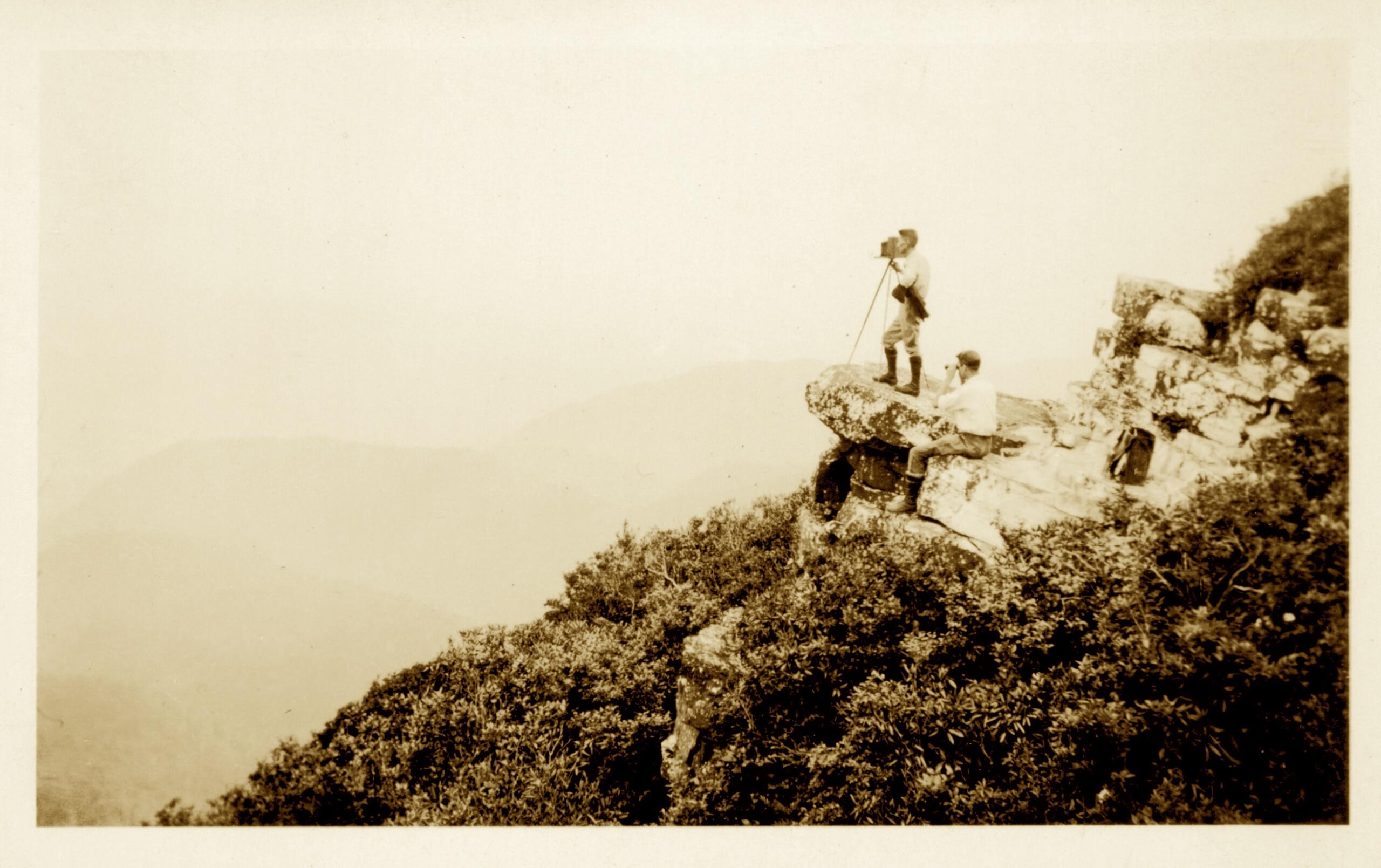 Two men on a rocky cliff using a vintage camera on a tripod, overlooking a foggy valley below.