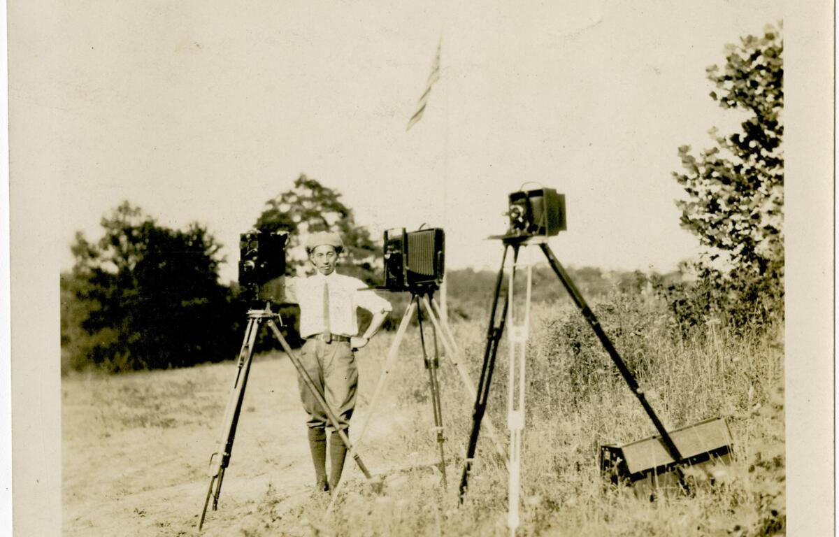 Man in a light shirt and cap stands in a grassy field beside three vintage cameras on tripods, outdoors with trees in the background.