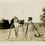 Man in a light shirt and cap stands in a grassy field beside three vintage cameras on tripods, outdoors with trees in the background.