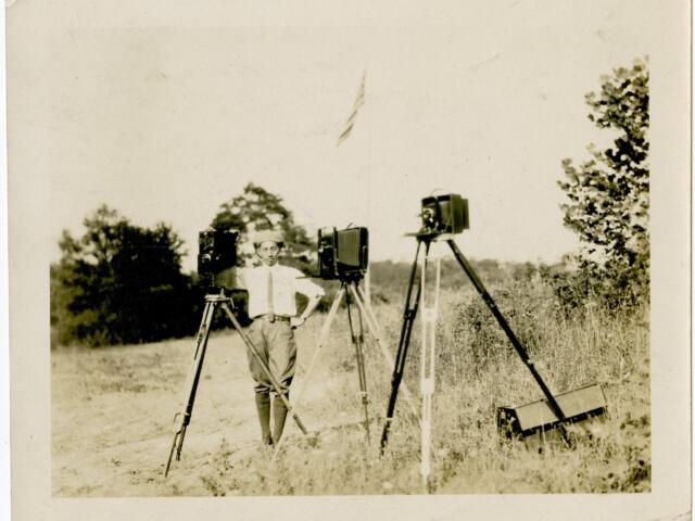 Man in a light shirt and cap stands in a grassy field beside three vintage cameras on tripods, outdoors with trees in the background.