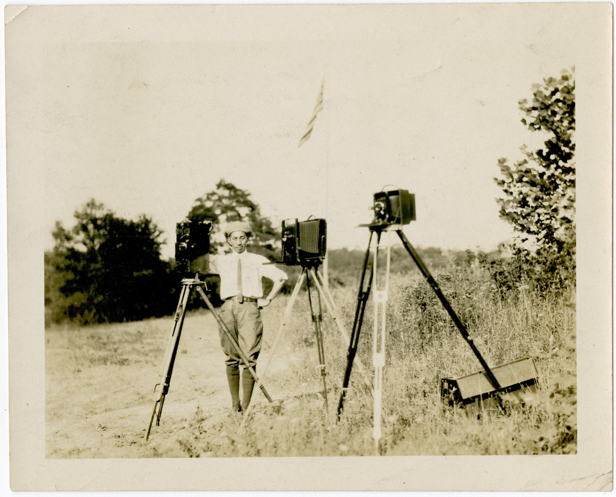 Man in a light shirt and cap stands in a grassy field beside three vintage cameras on tripods, outdoors with trees in the background.
