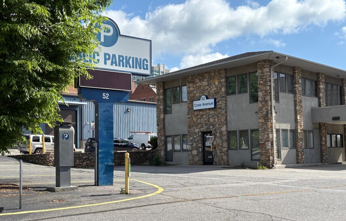 Public parking sign and stone office building at 52 Coxe Avenue, with a tree-lined foreground and a blue sky.