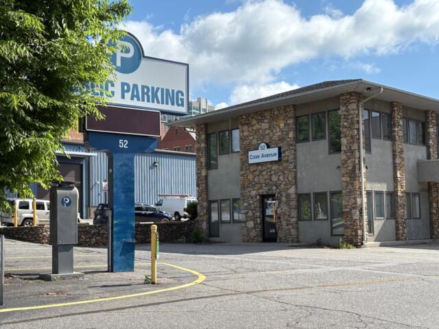 Public parking sign and stone office building at 52 Coxe Avenue, with a tree-lined foreground and a blue sky.