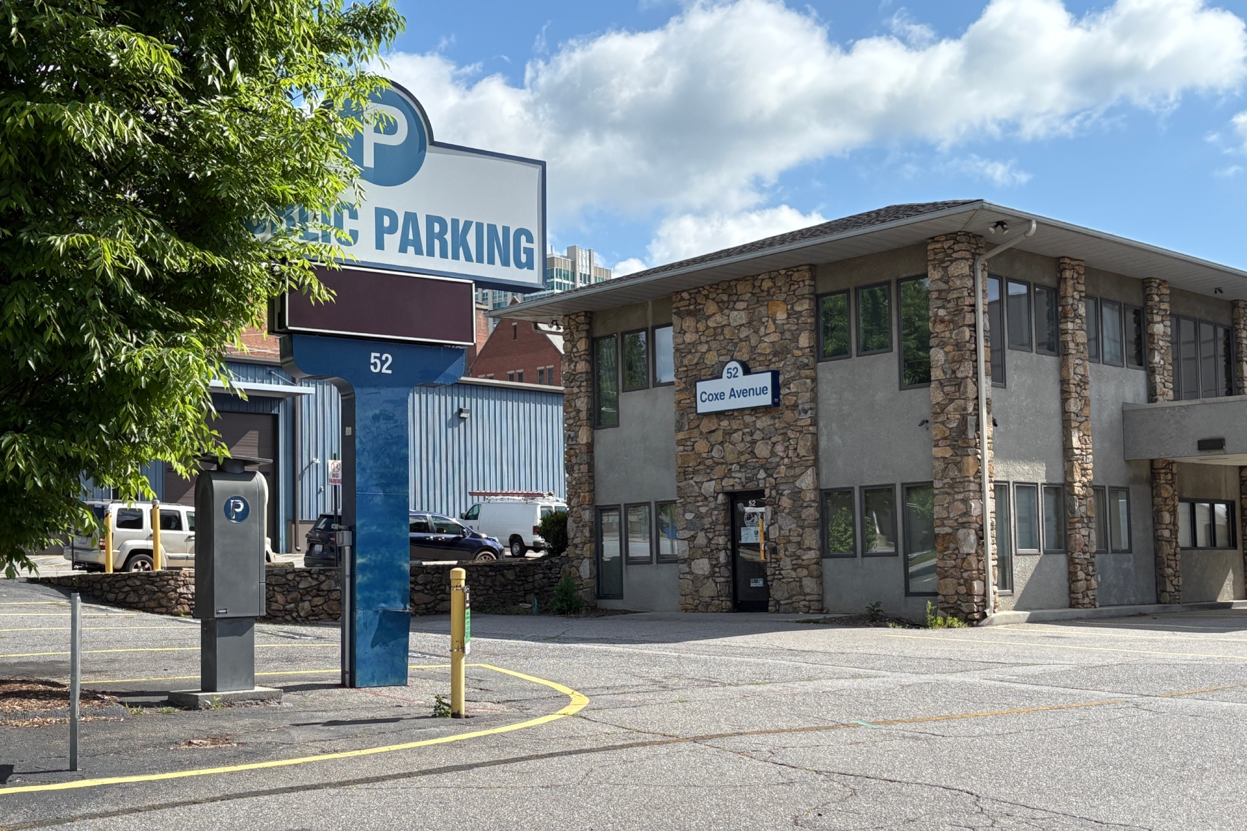 Public parking sign and stone office building at 52 Coxe Avenue, with a tree-lined foreground and a blue sky.