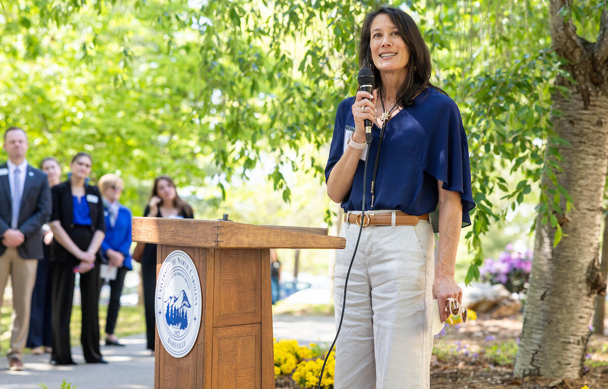 Woman in a blue blouse speaks into a microphone at a wooden podium with a university seal, outdoors.