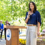 Woman in a blue blouse speaks into a microphone at a wooden podium with a university seal, outdoors.