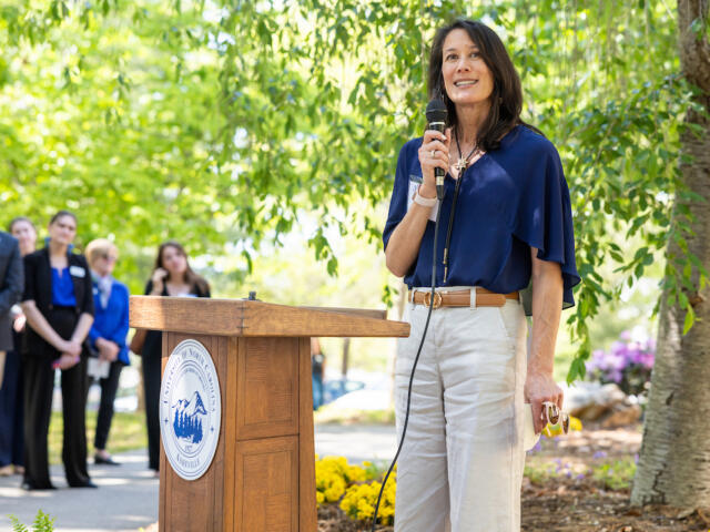 Woman in a blue blouse speaks into a microphone at a wooden podium with a university seal, outdoors.