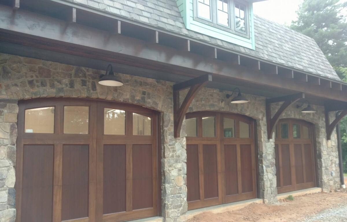 Four wooden arched garage doors set in a stone building, with a covered porch and rustic stone columns. Outdoor lamps hang from the eave above the doors, and a shingled roof lines the upper level.
