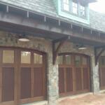 Four wooden arched garage doors set in a stone building, with a covered porch and rustic stone columns. Outdoor lamps hang from the eave above the doors, and a shingled roof lines the upper level.