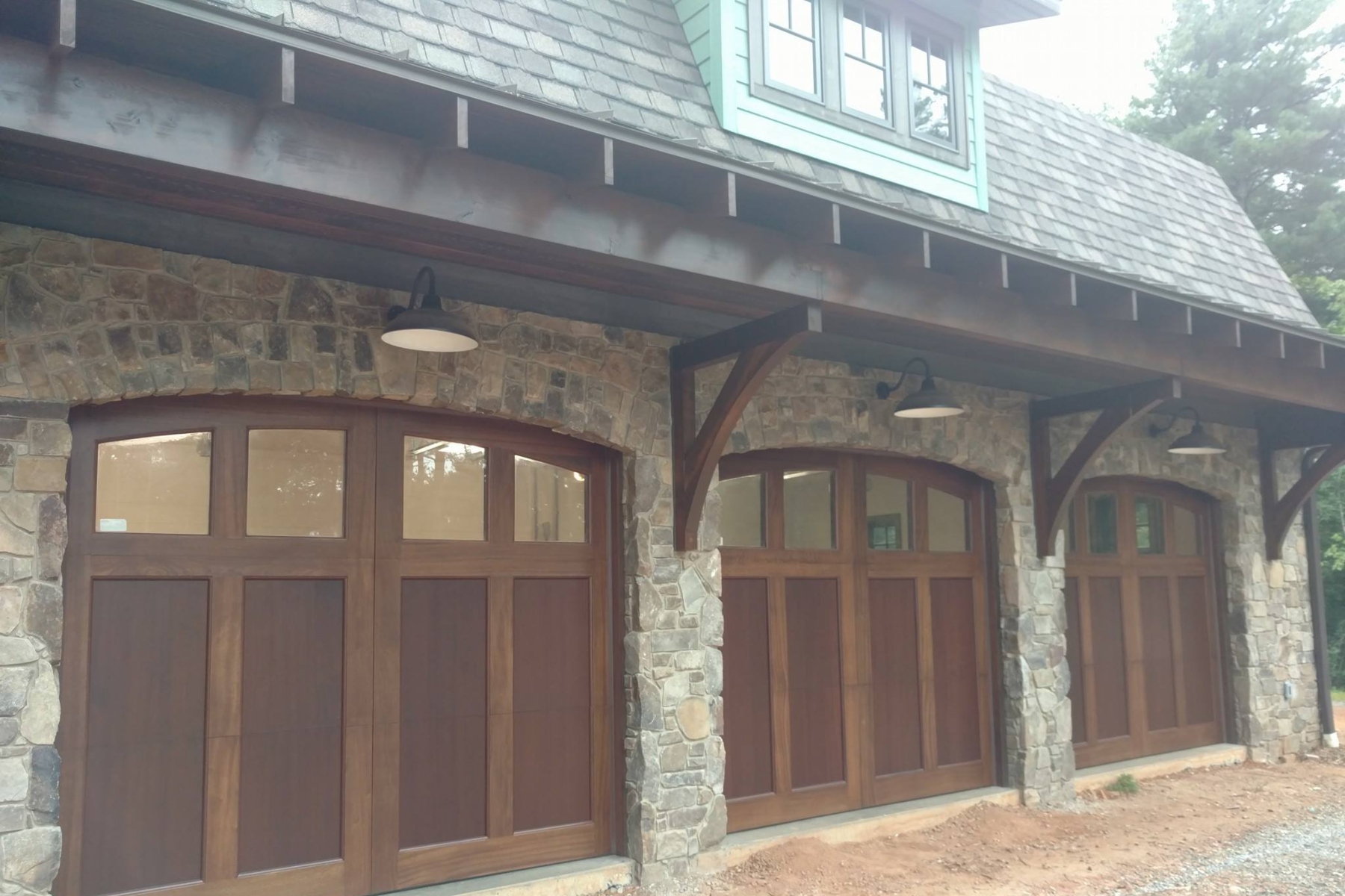 Four wooden arched garage doors set in a stone building, with a covered porch and rustic stone columns. Outdoor lamps hang from the eave above the doors, and a shingled roof lines the upper level.