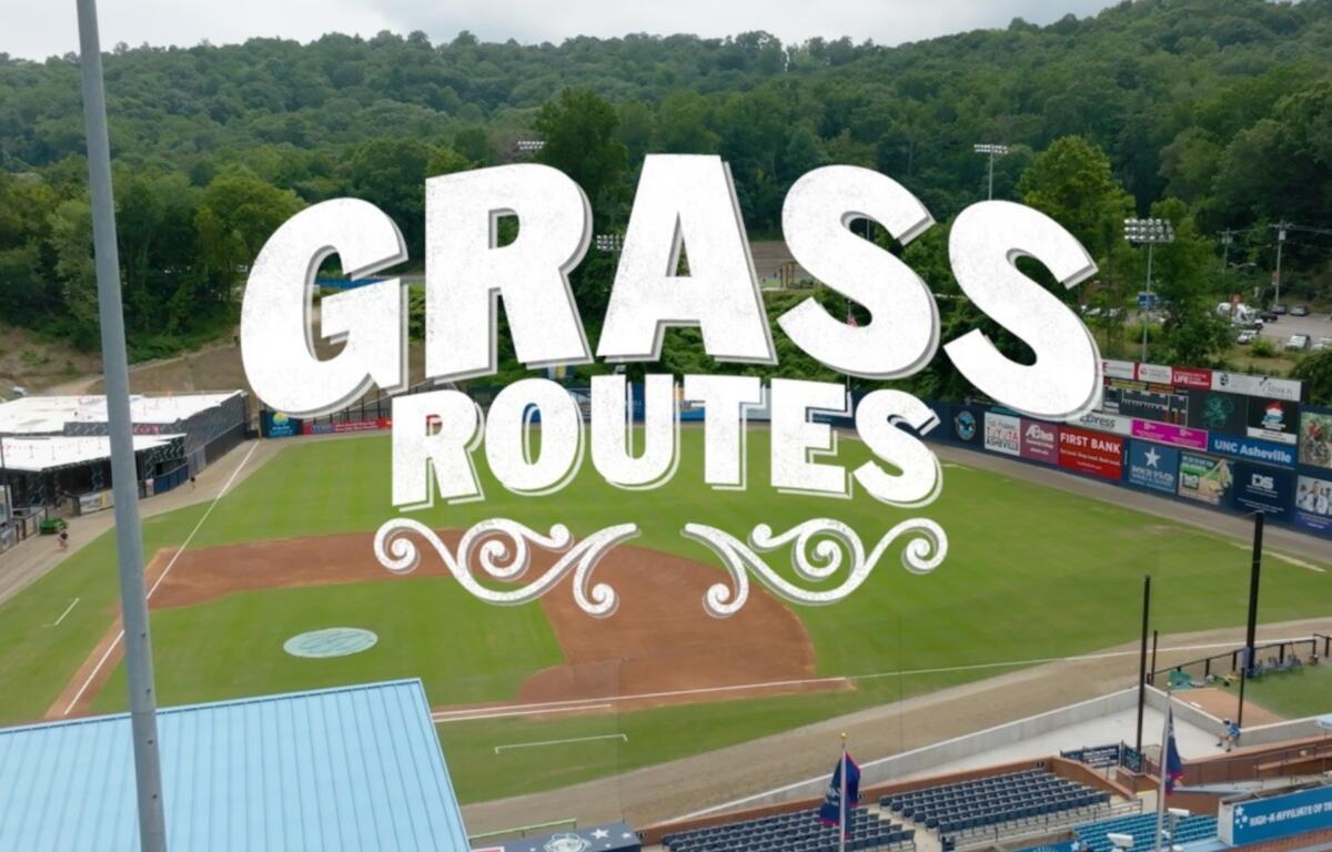 Aerial view of a baseball field with a bold white 'GRASS ROUTES' graphic overlay across the infield and stands.