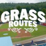 Aerial view of a baseball field with a bold white 'GRASS ROUTES' graphic overlay across the infield and stands.