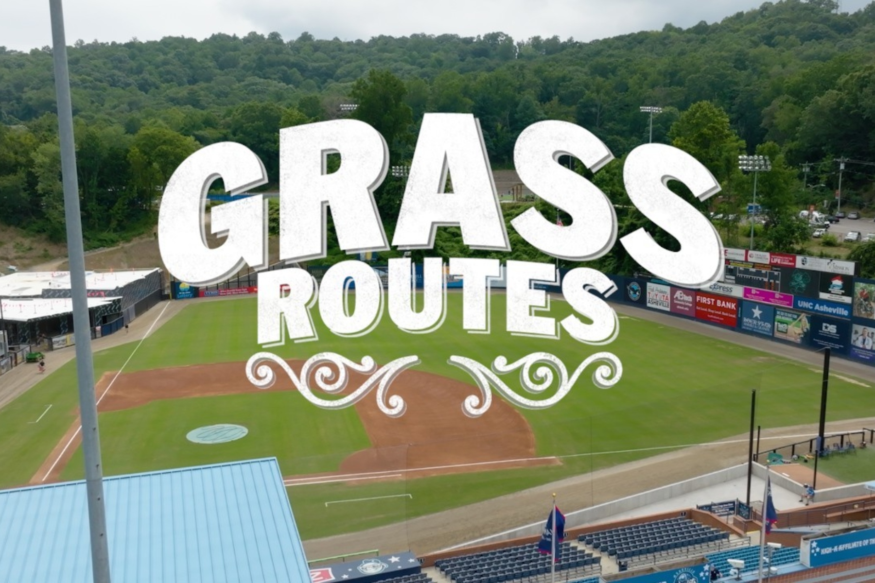 Aerial view of a baseball field with a bold white 'GRASS ROUTES' graphic overlay across the infield and stands.