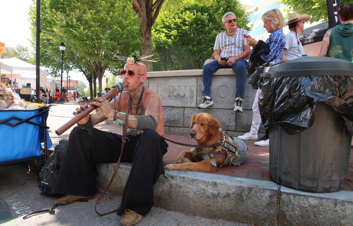 Tattoos musician with a flower headpiece plays a didgeridoo on a city sidewalk, a golden retriever in a vest rests nearby.