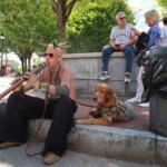 Tattoos musician with a flower headpiece plays a didgeridoo on a city sidewalk, a golden retriever in a vest rests nearby.
