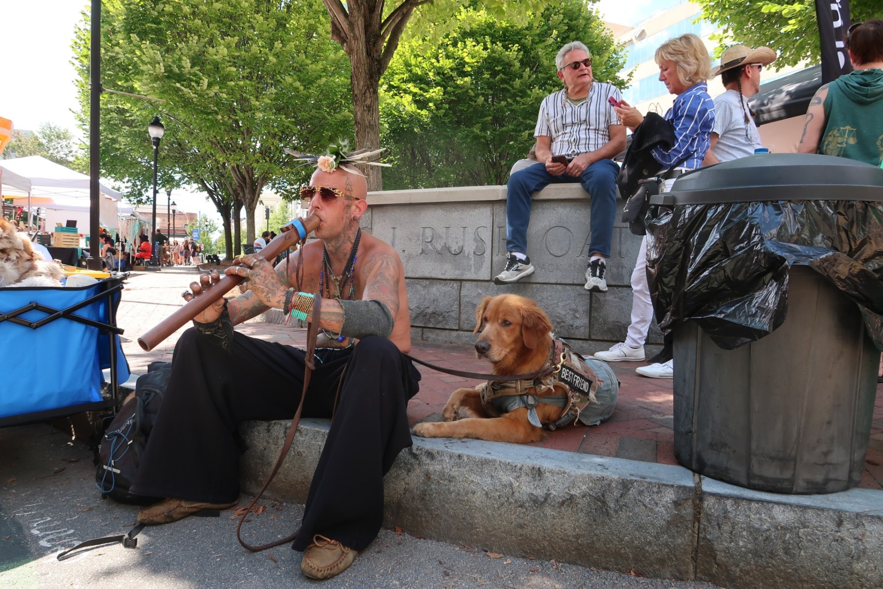 Tattoos musician with a flower headpiece plays a didgeridoo on a city sidewalk, a golden retriever in a vest rests nearby.