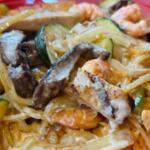 Creamy pasta dish with shrimp, sliced chicken, onions, zucchini, and other vegetables on a pink plate.