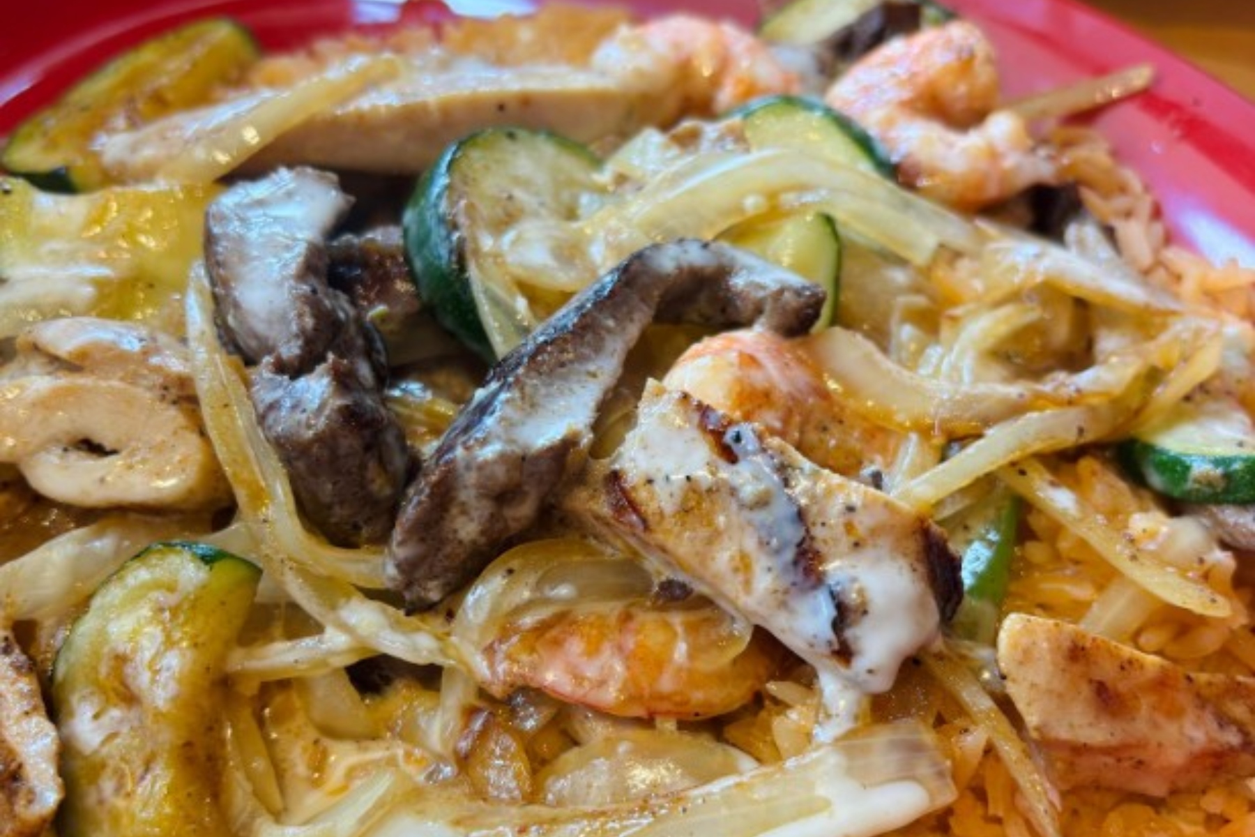Creamy pasta dish with shrimp, sliced chicken, onions, zucchini, and other vegetables on a pink plate.