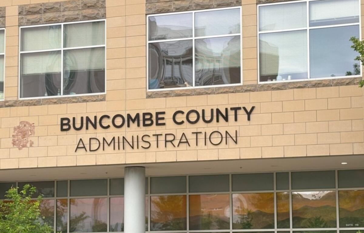 Front view of Buncombe County Administration building with beige brick and large windows; a decorative emblem is visible on the left side of the facade.