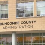 Front view of Buncombe County Administration building with beige brick and large windows; a decorative emblem is visible on the left side of the facade.