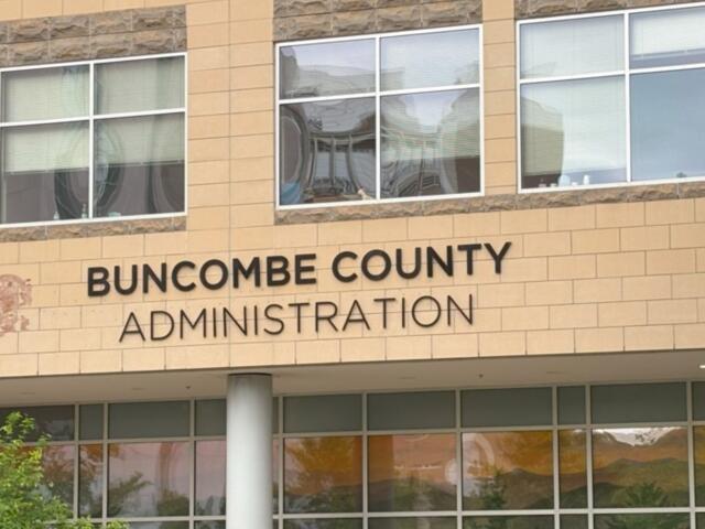 Front view of Buncombe County Administration building with beige brick and large windows; a decorative emblem is visible on the left side of the facade.
