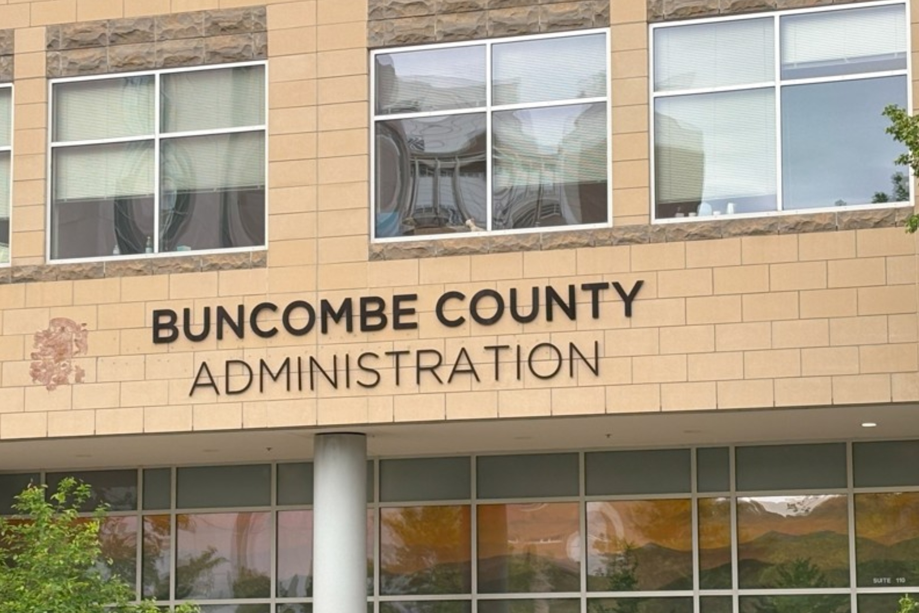 Front view of Buncombe County Administration building with beige brick and large windows; a decorative emblem is visible on the left side of the facade.
