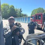 Group of firefighters outside a row of blue storage units; a man in uniform holds a black-and-tan dog, while a red fire truck is parked in the background.
