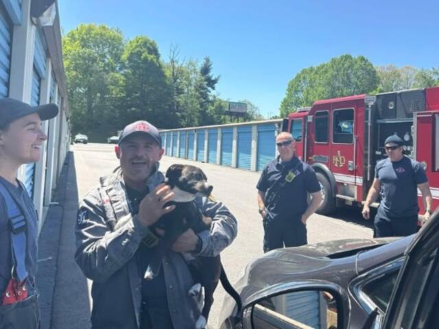 Group of firefighters outside a row of blue storage units; a man in uniform holds a black-and-tan dog, while a red fire truck is parked in the background.