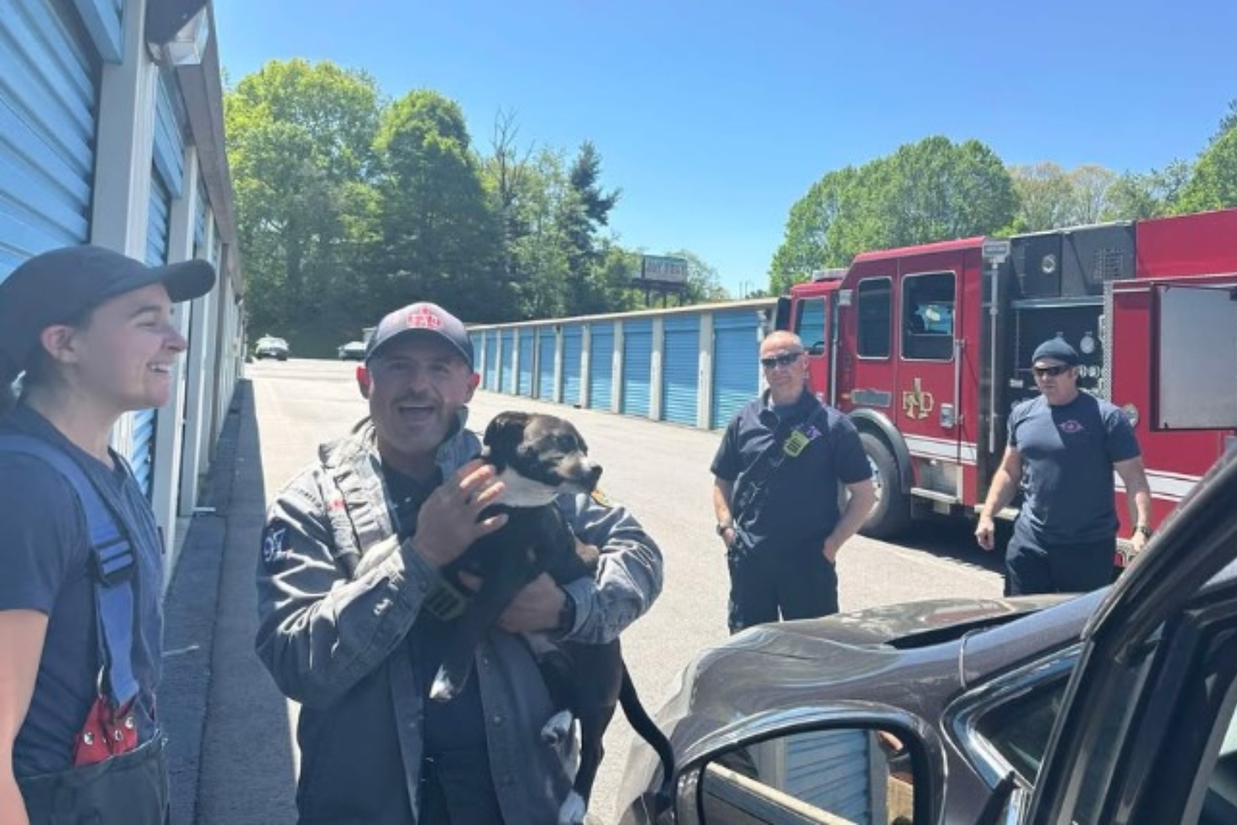 Group of firefighters outside a row of blue storage units; a man in uniform holds a black-and-tan dog, while a red fire truck is parked in the background.