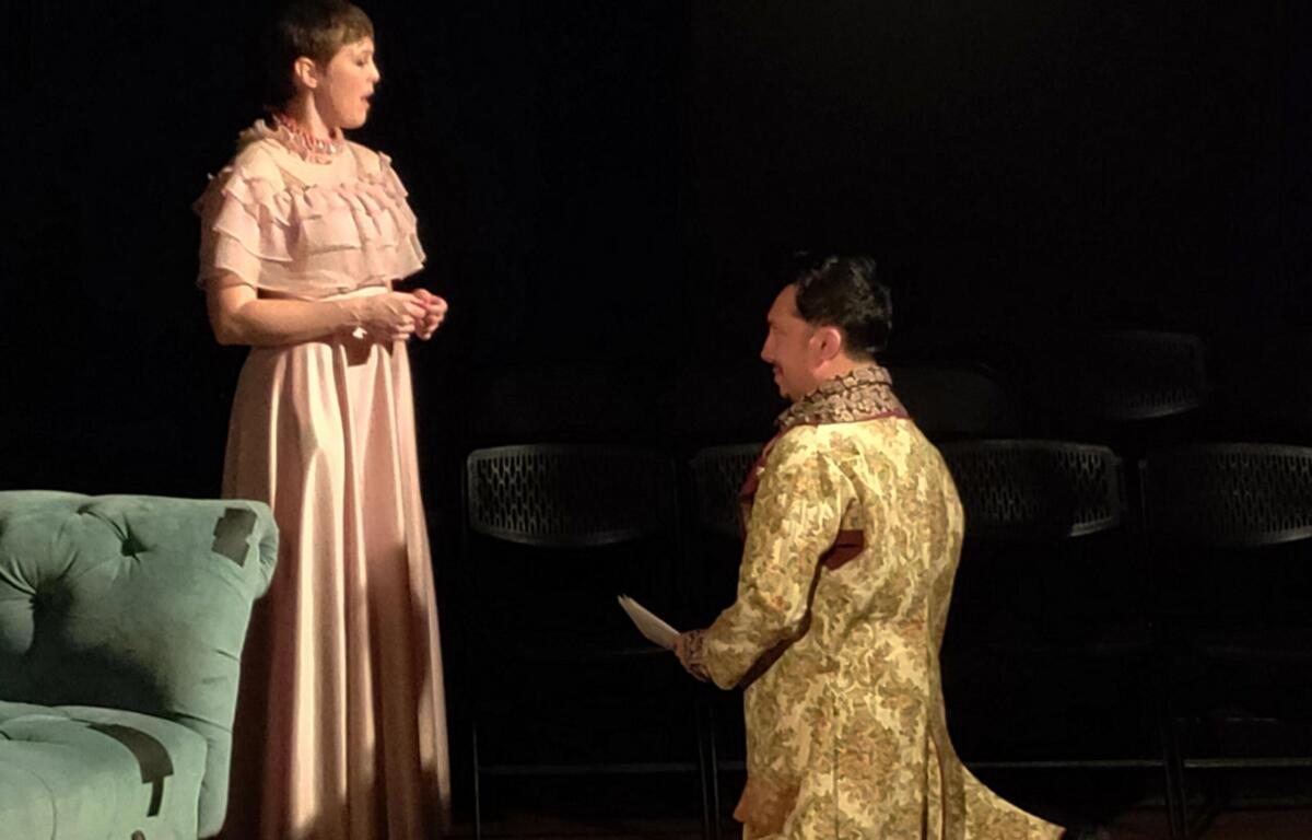 Stage scene with two performers in historical costumes: a standing woman in a pink, ruffled dress and a kneeling person in a floral gown, on a dark backdrop with a green couch visible to the left.