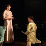 Stage scene with two performers in historical costumes: a standing woman in a pink, ruffled dress and a kneeling person in a floral gown, on a dark backdrop with a green couch visible to the left.