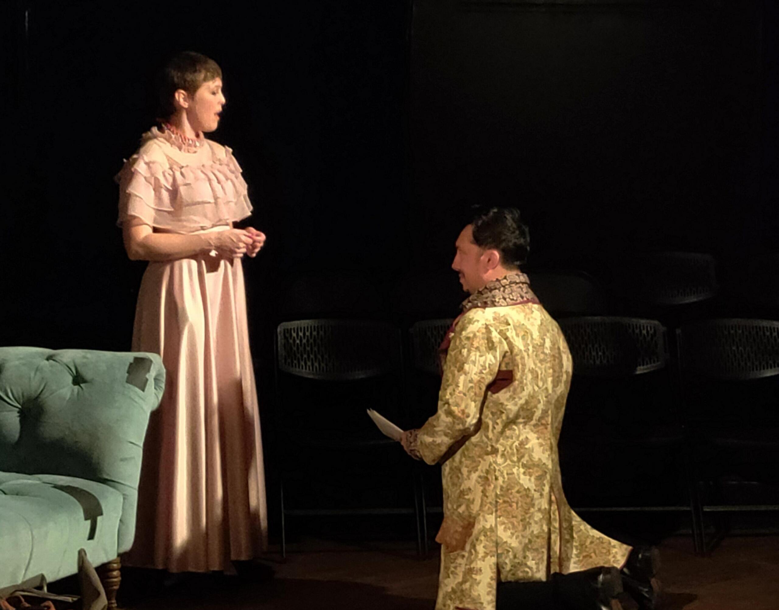 Stage scene with two performers in historical costumes: a standing woman in a pink, ruffled dress and a kneeling person in a floral gown, on a dark backdrop with a green couch visible to the left.