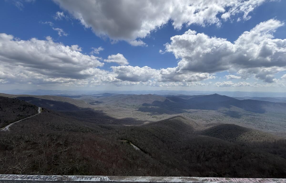 Expansive view of rolling mountains under a blue, cloud-filled sky from a lookout railing in the foreground.