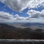 Expansive view of rolling mountains under a blue, cloud-filled sky from a lookout railing in the foreground.