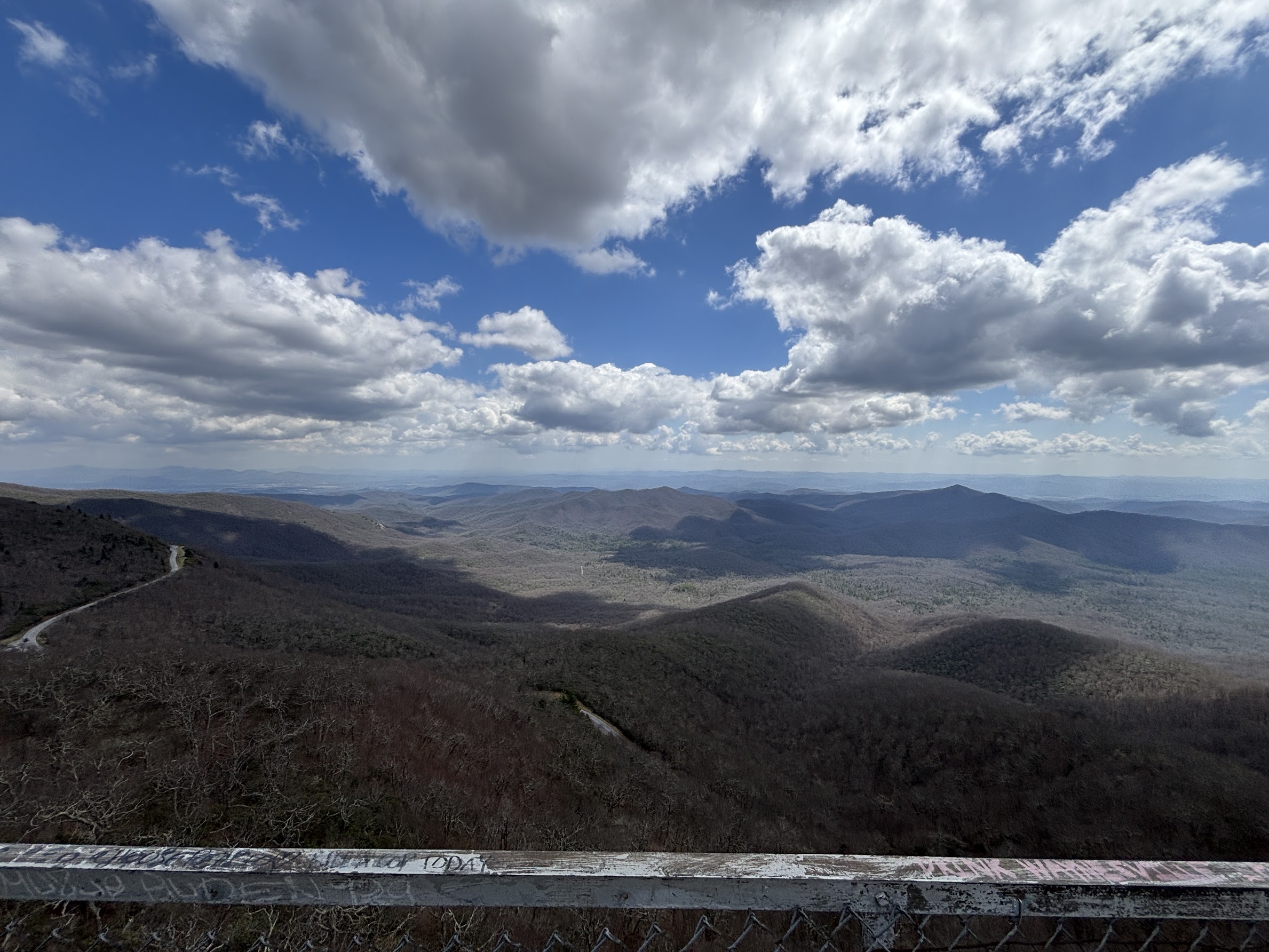 Expansive view of rolling mountains under a blue, cloud-filled sky from a lookout railing in the foreground.