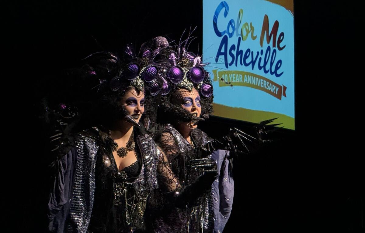 Two performers in glittery black costumes with large purple headdresses on stage; a Color Me Asheville banner is in the background.