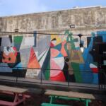 Colorful geometric mural on a brick wall with 'Coffee and Doughnuts' lettering, outdoor seating and string lights in front.