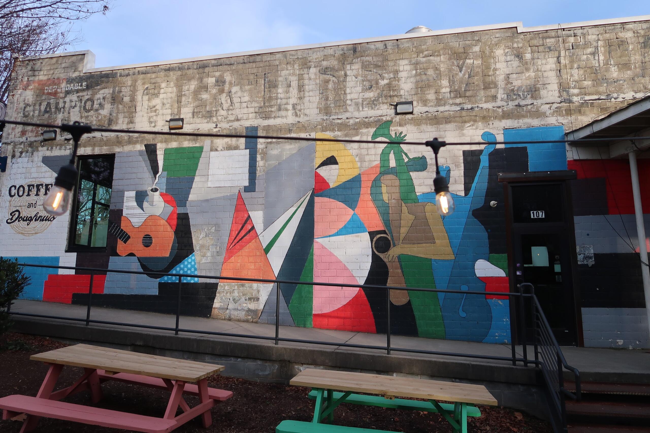 Colorful geometric mural on a brick wall with 'Coffee and Doughnuts' lettering, outdoor seating and string lights in front.