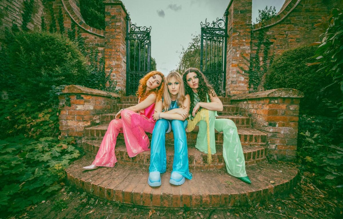 Three women in colorful outfits sit on brick steps in front of an ornate wrought-iron gate with ivy-covered brick walls.