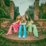 Three women in colorful outfits sit on brick steps in front of an ornate wrought-iron gate with ivy-covered brick walls.