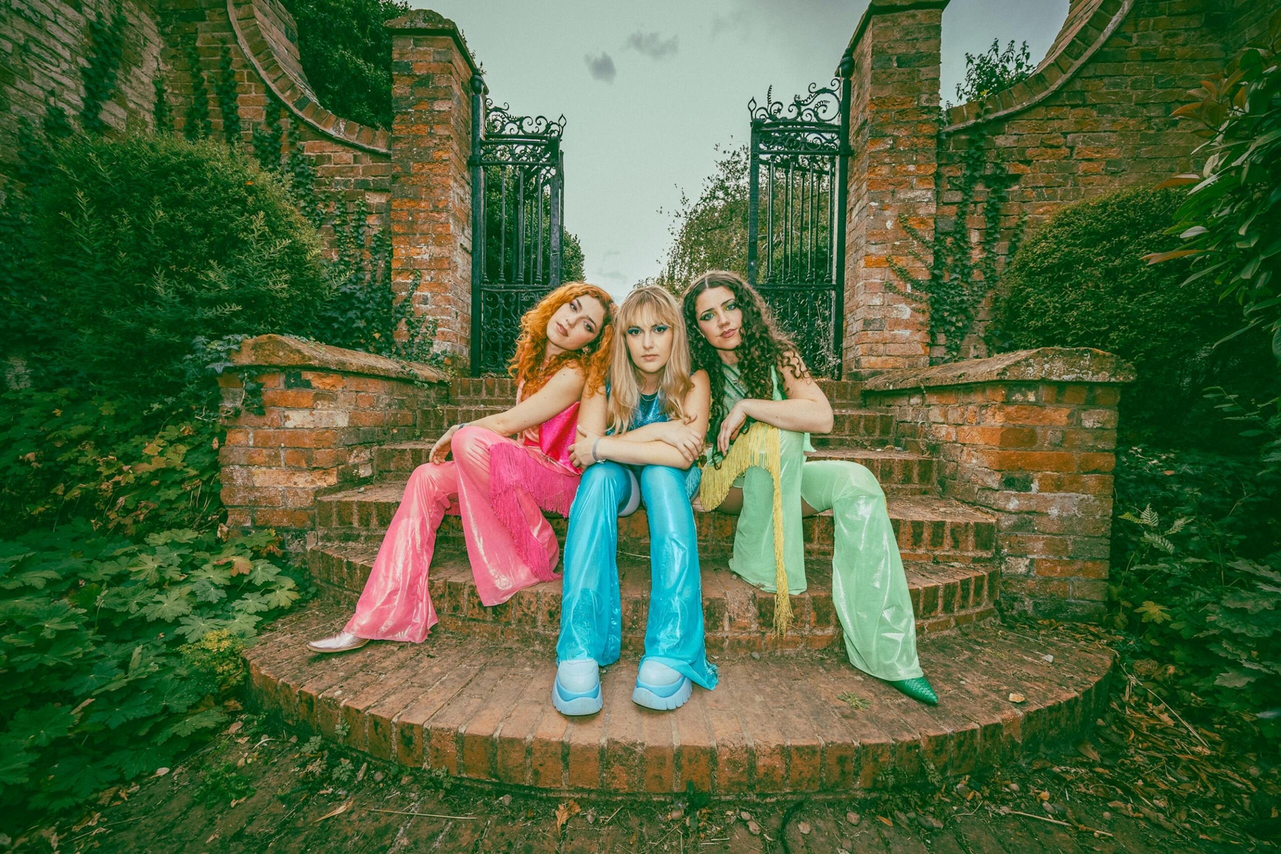 Three women in colorful outfits sit on brick steps in front of an ornate wrought-iron gate with ivy-covered brick walls.