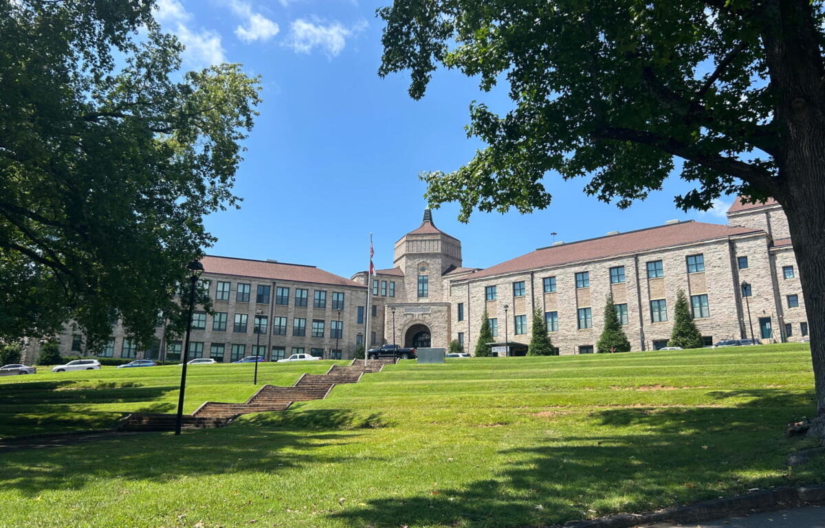 Stone campus building with central arched entrance, flagpole, and surrounding green lawn on a sunny day; trees frame the scene.