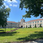 Stone campus building with central arched entrance, flagpole, and surrounding green lawn on a sunny day; trees frame the scene.