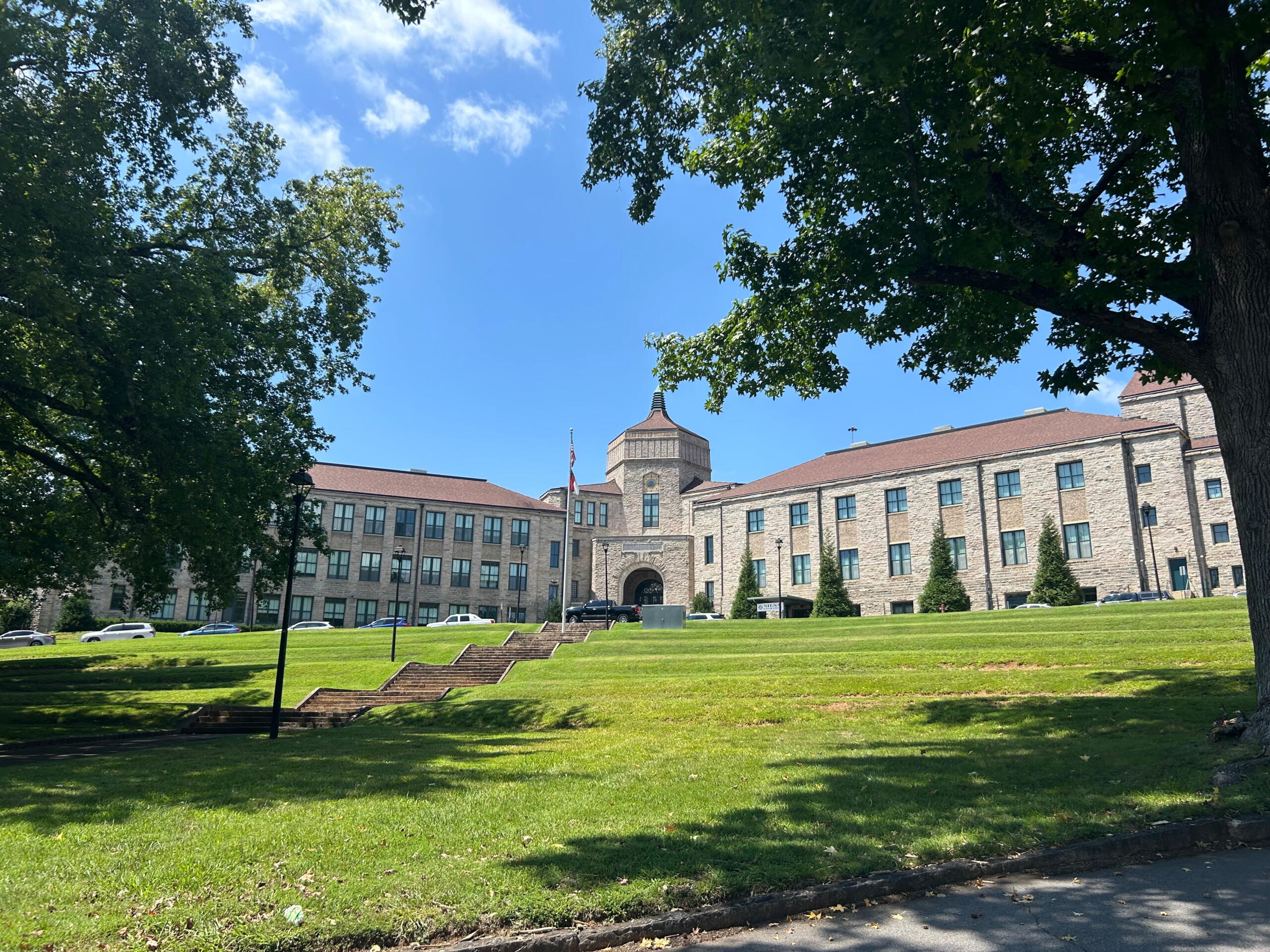 Stone campus building with central arched entrance, flagpole, and surrounding green lawn on a sunny day; trees frame the scene.